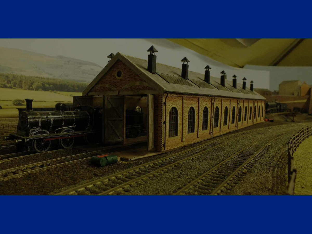 "Two kits were used with smoke stacks, (from kit offcuts and plastikard roofs mounted on Peco track spikes), added rooflights and heavier gables.
The downpipes use florist's wire and guttering is from corn flake packet card.
Internal walls use yellow brick paper on thicker card for added thickness.
The end walls were altered with the addition of saving brick arches and concrete lintels.
The doors have added plastic strip on the outside for thickness and thicker bracing on the insides.
Finally the roof was slated with Superquick slate paper cut into strips and hung using the etched slates as guides.
The brickwork was coloured using pastel pencils - laborious but worthwhile!"